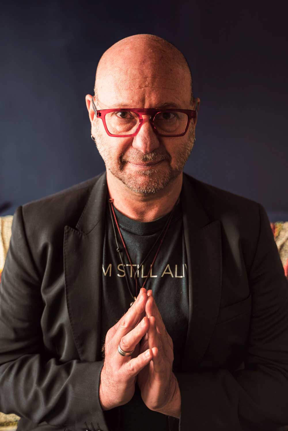 A portrait of Murray Nossel in a blazer and t-shirt that reads "I'm still alive" with his hands clasped